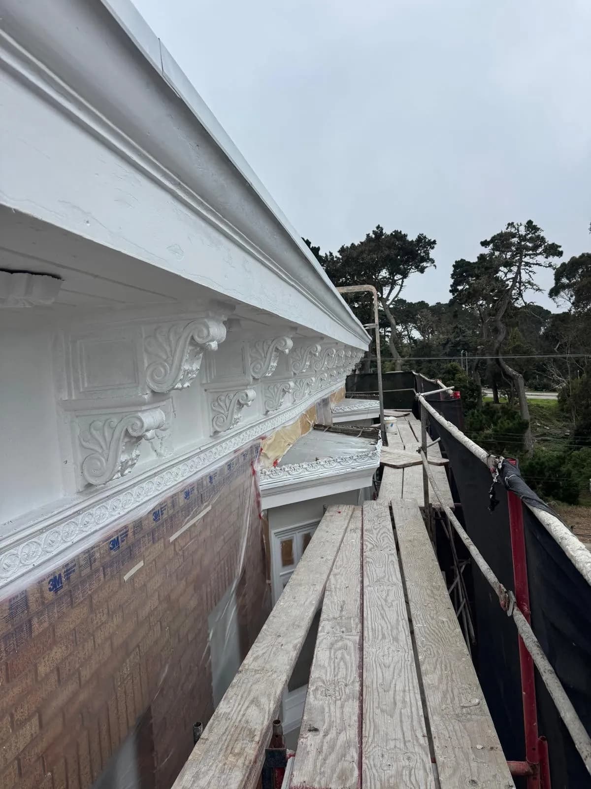 White cornice brackets and dentil molding seen from wooden scaffold planks, with masking film on the wall below