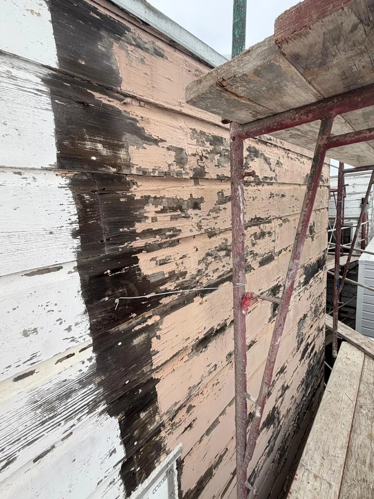 Wood siding with paint scraped and peeled away to bare wood, next to a scaffold frame