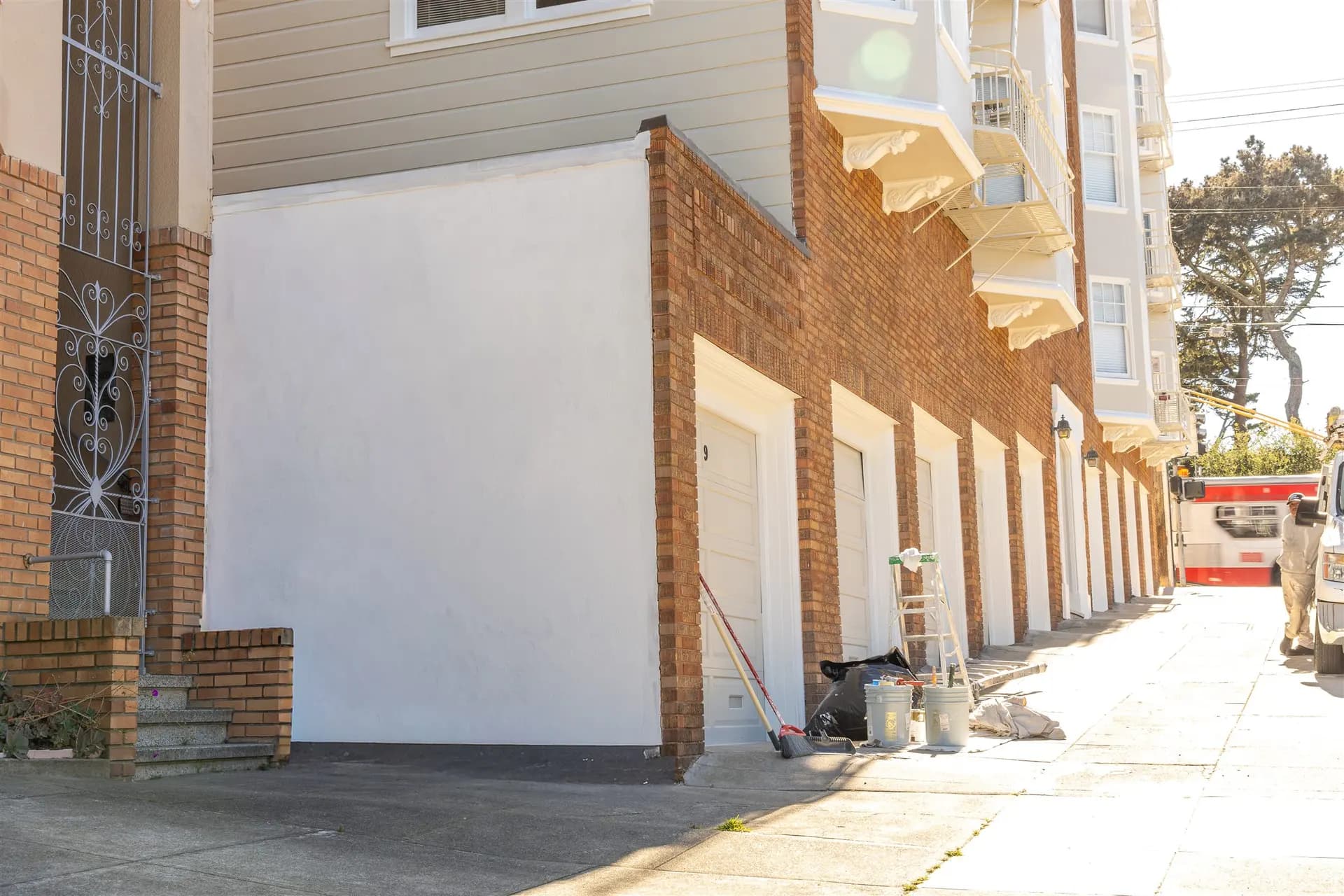 Side of the San Francisco brick apartment building with a white wall and white garage doors, a ladder and paint buckets still on the sidewalk